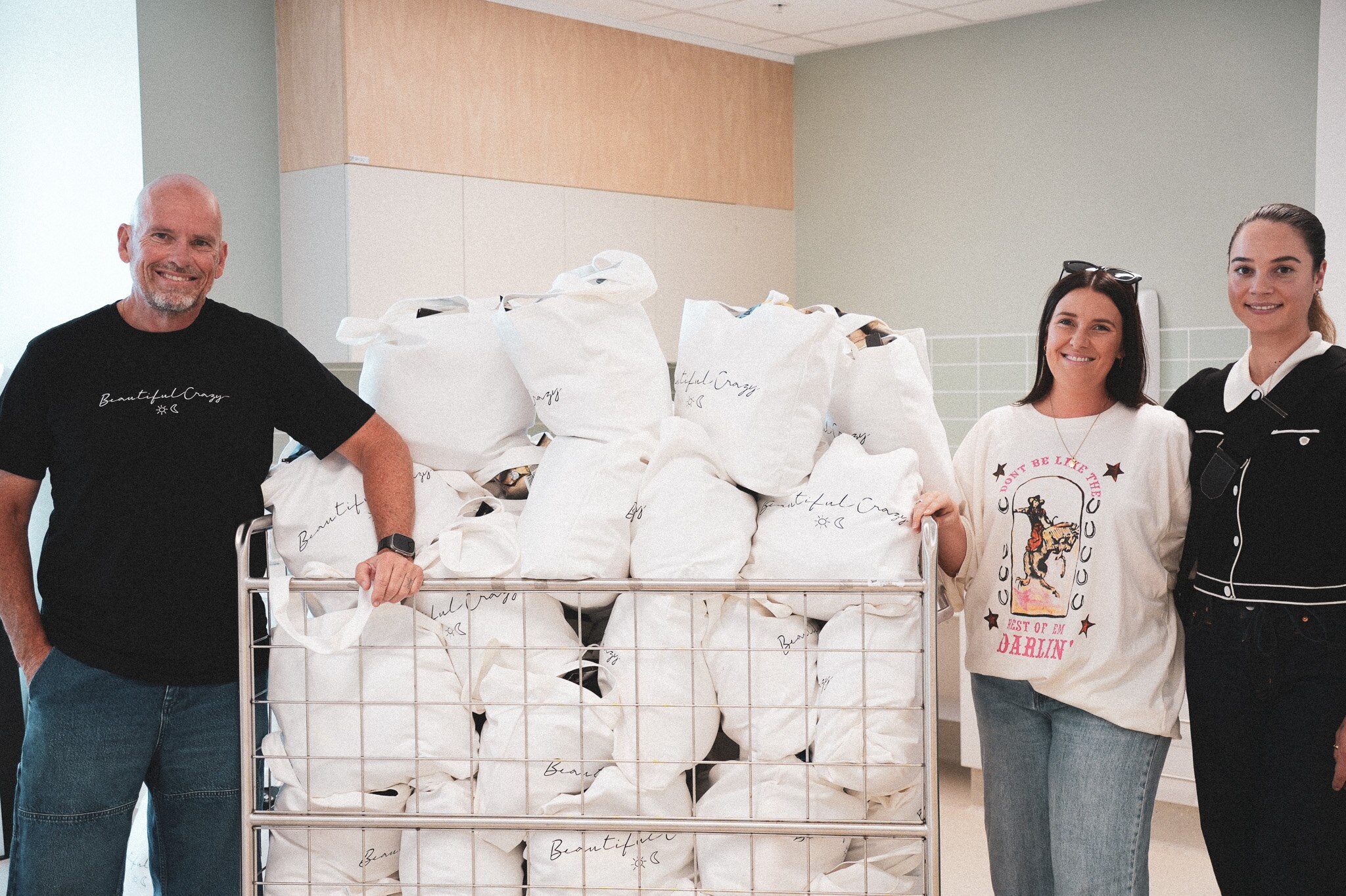 A man and two women standing next to a cart full of care packages inside a hospital ward.