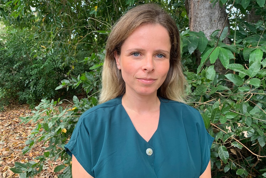 A close-up portrait of a woman standing in front of some trees, smiling slightly.