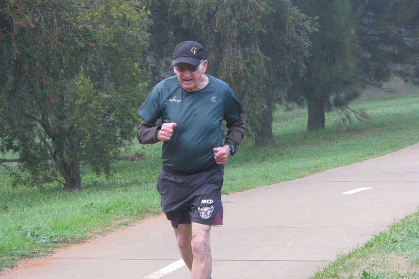 An 83-year-old man pumping his arms as he runs on a path bordered by trees