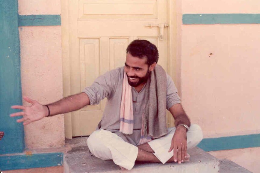 A young Narendra Modi sits on a door step, smiling while extending a hand out