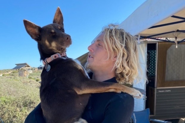 A young man with blonde hair holds a dog in his arms, in a bush area.