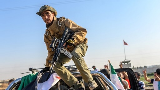 A soldier in military gear climbs on top of a car with his gun, surrounded by cheering people and flags