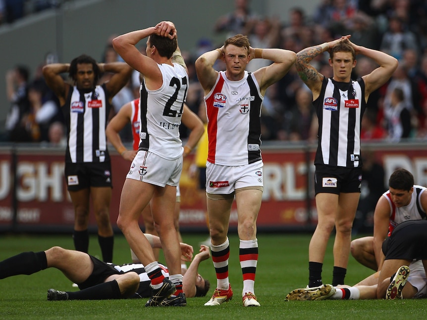 A group of AFL footballers stand with hands on heads or lie on the ground exhausted after a drawn in grand final.