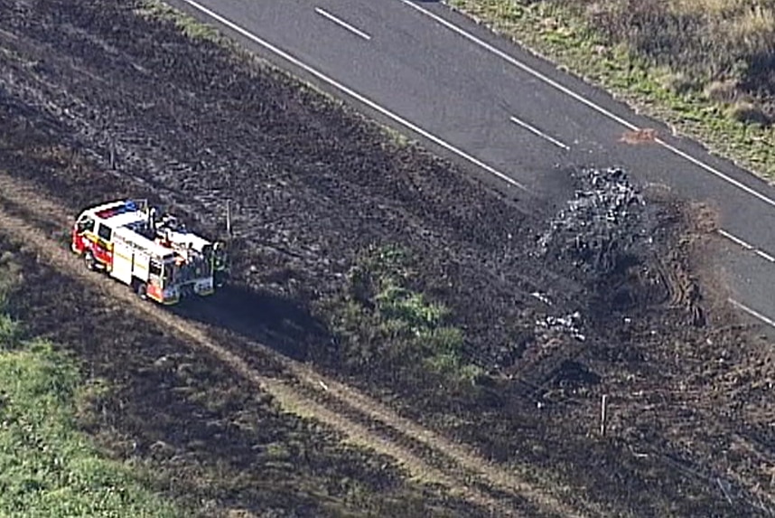 A fire truck drives past strewn car parts and dirt and the charred grass from a fiery fatal crash.