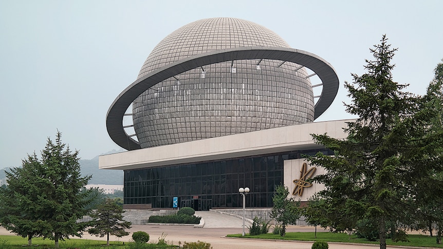 Colour photograph of a low level but long building with a large grey dome structure similar to a planet in shape, on it's roof.