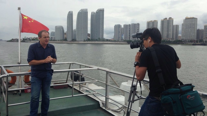 A man stands on a boat with a Chinese flag and highrise in the background while another man looks through a video camera