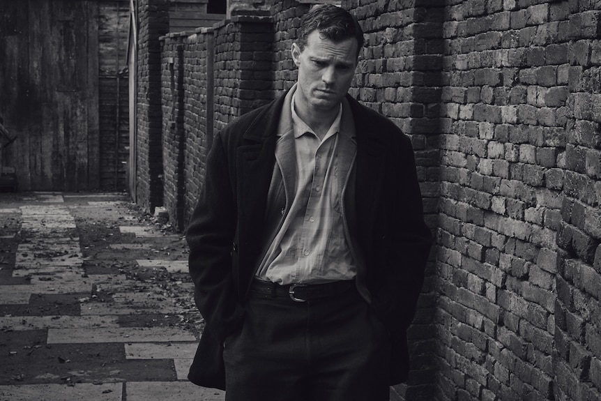 A black and white image of a serious middle-aged man with his head bowed and hands in his pockets, walking along the street