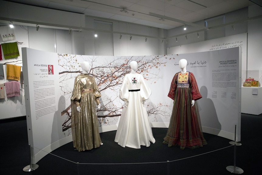 Three dresses in traditional Afghan embroidery on display in an exhibition, in front of a wall of text about each piece.