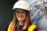 Felicity Furey, wearing a hard hat and high-vis vest, stands in an office in front of an image of the West Gate Tunnel.