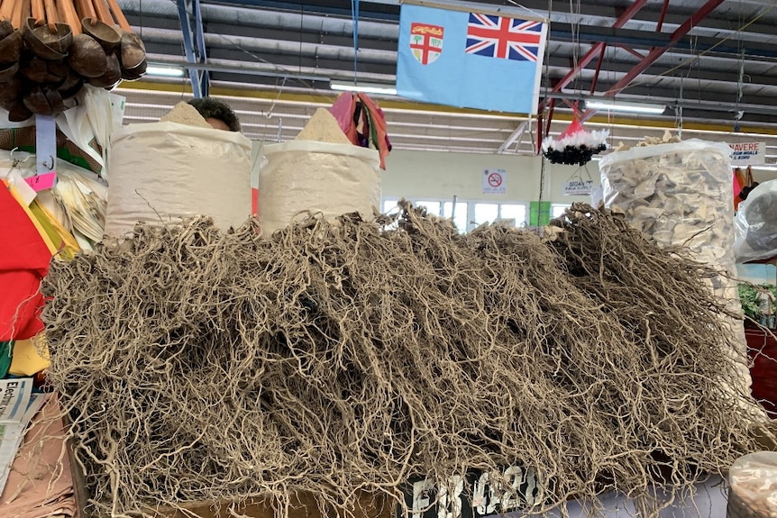 A table full of twisted dry grassy roots on a table in a market with a Fijian flag in the background.