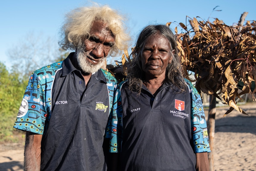 An older Indigenous couple, man with a mop of grey hair and beard, stand together under blue sky, sand.