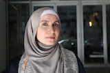 A woman in a hijab stands in front of a glass wall in the afternoon light.