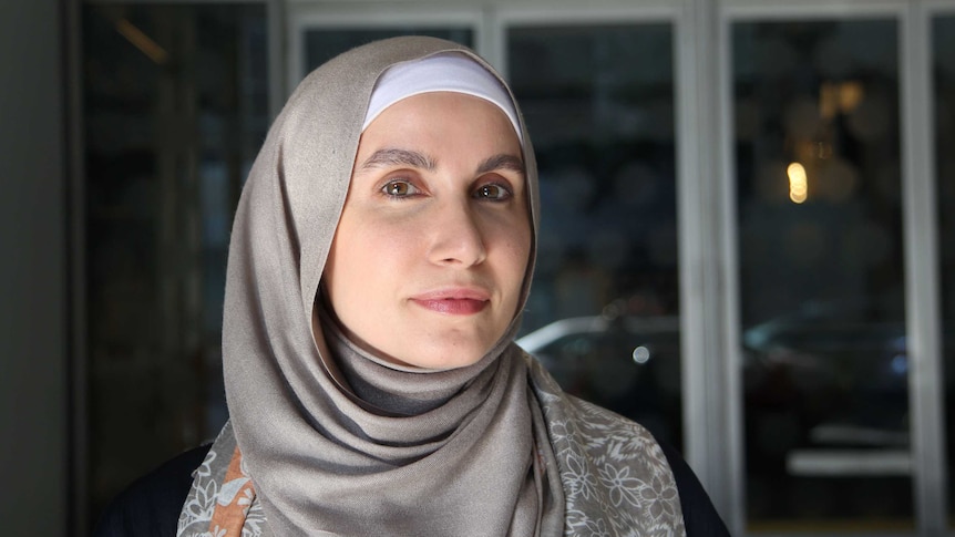 A woman in a hijab stands in front of a glass wall in the afternoon light.