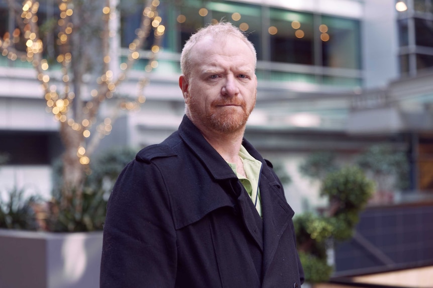 A close up portrait of a man in front of a nondescript blurred urban background.