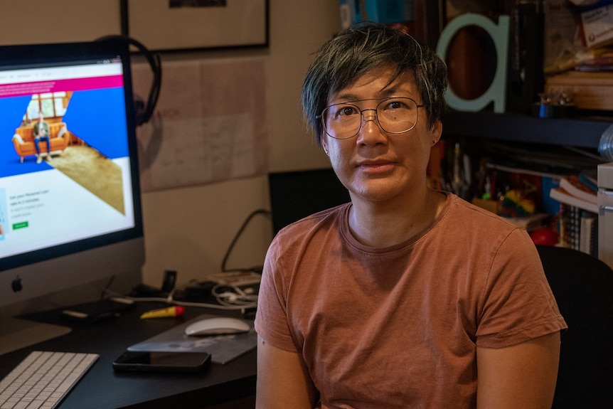 A middle aged Asian woman with short dark hair, glasses and tanned skin sits at a desk in front of a computer.