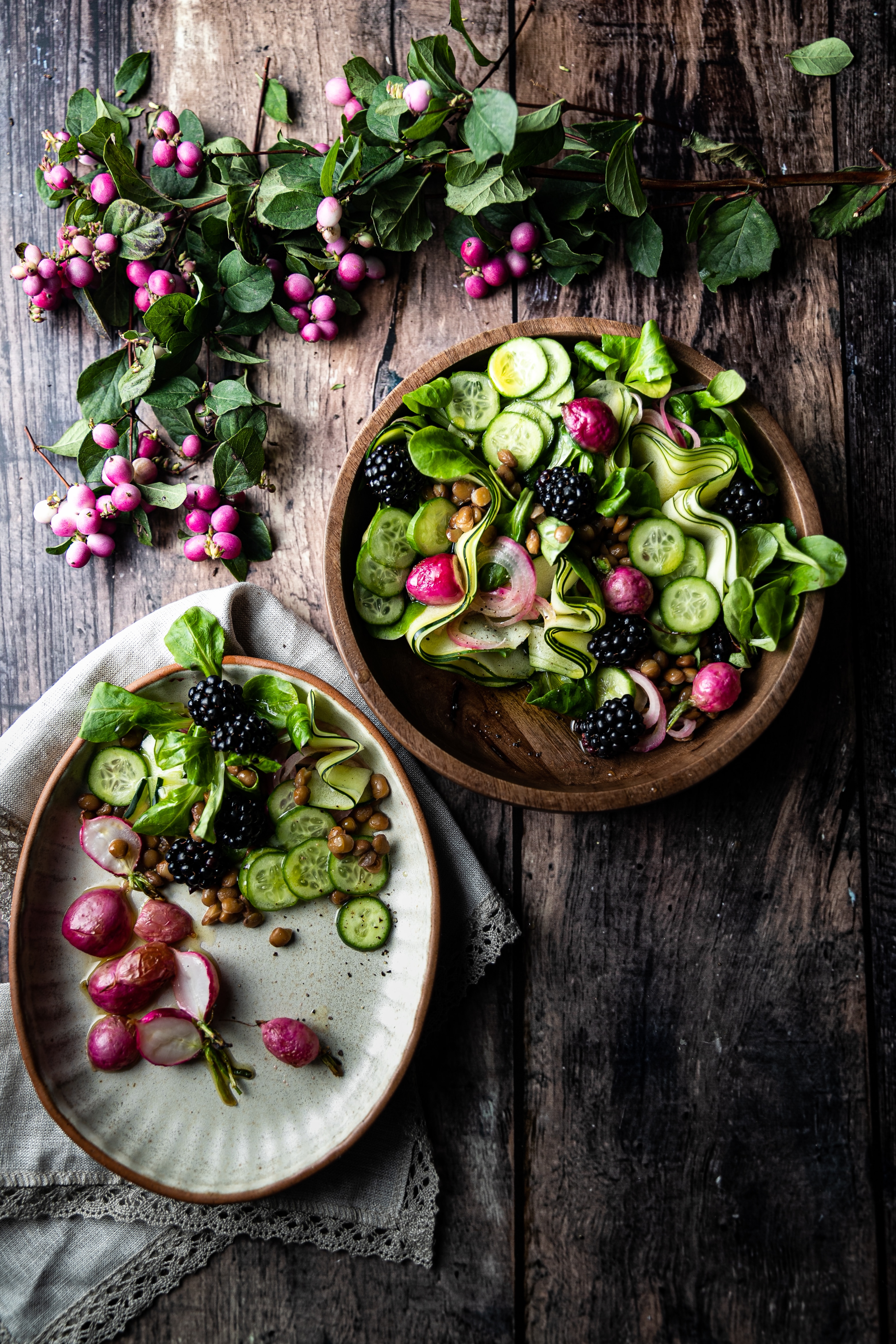 Two rustic bowls hold green salads, punctuated by the pink shades of radishes and black of mulberries. 