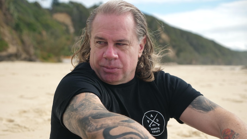A  tattooed man with shoulder length blond hair sits on the beach, looking down, board behind him.