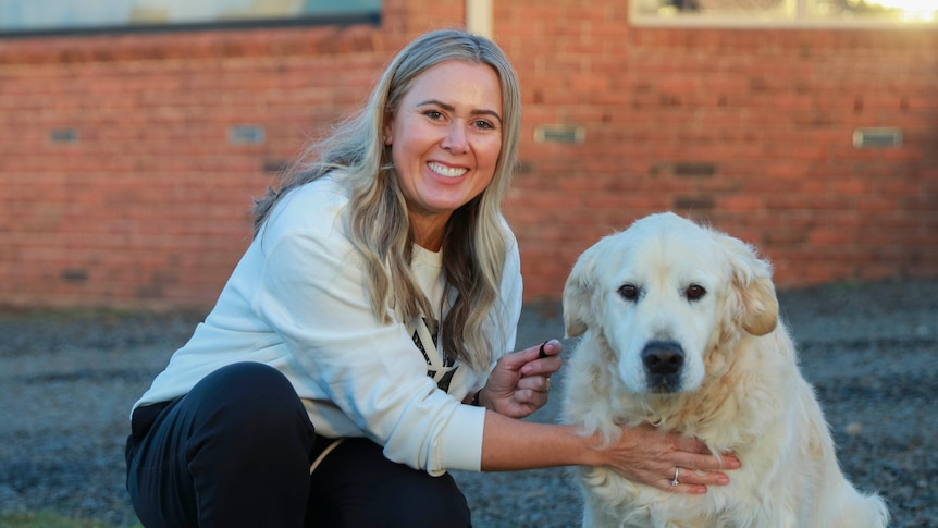 A blonde-haired young woman kneels next to a golden retriever dog