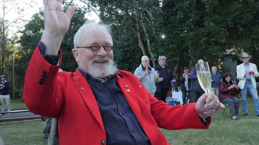 Betrand Cadart in red jacket sits in a chair with glass of champagne, smiling and waving