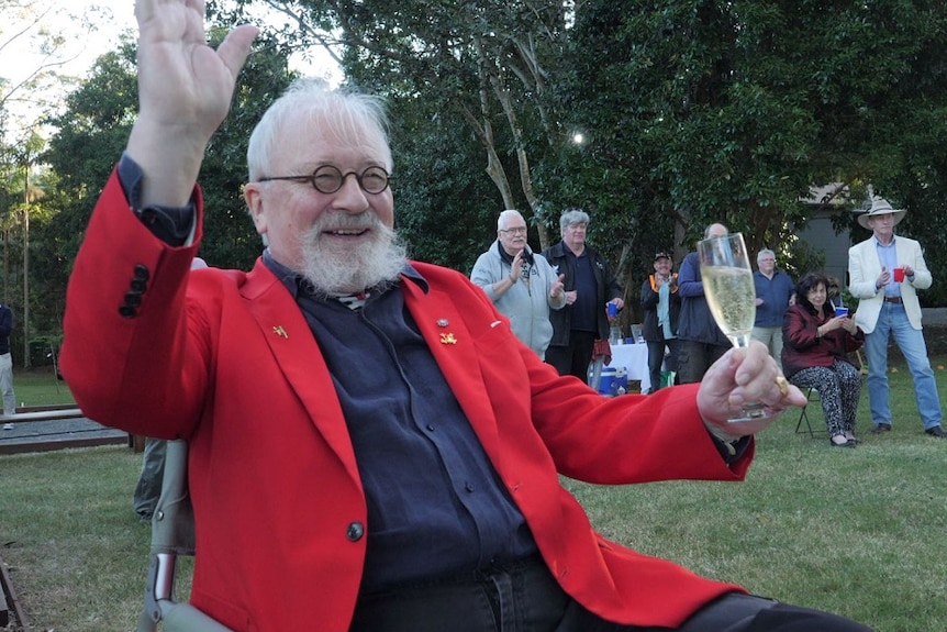 Betrand Cadart in red jacket sits in a chair with glass of champagne, smiling and waving.