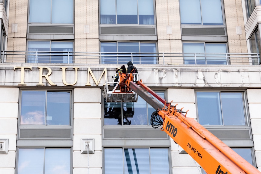 Two men remove the letters T.R.U.M.P one by one from the side of a building.