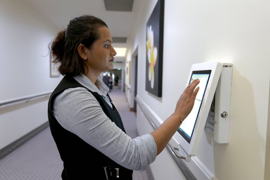 A nurse in a collared shirt stands in the hallway of an aged care home touching a screen on the wall.