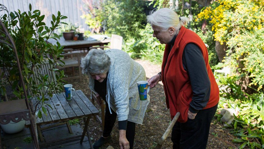 Kristin brushes leaves off a garden chair as Hilary holds a cup of coffee.