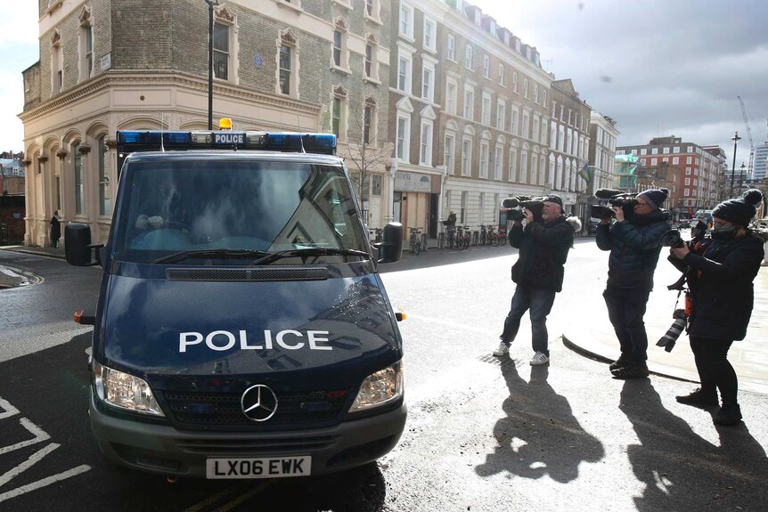 A police van drives past three camera operators as it turns into a court building off a London street.