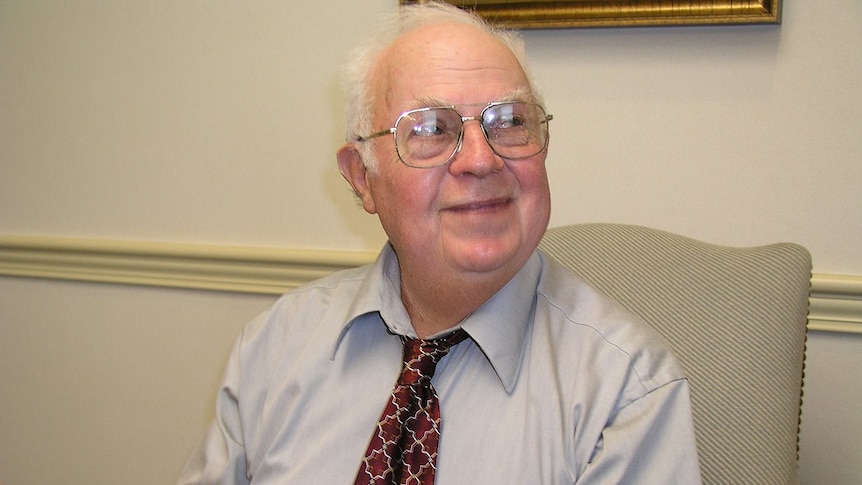 A friendly-looking older man in a business shirt and tie sits at a table, looking to the right of the frame.