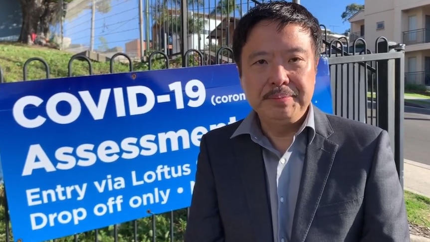 A man in a grey suit and open-necked blue business shirt stands in front of a COVID-19 assessment clinic sign