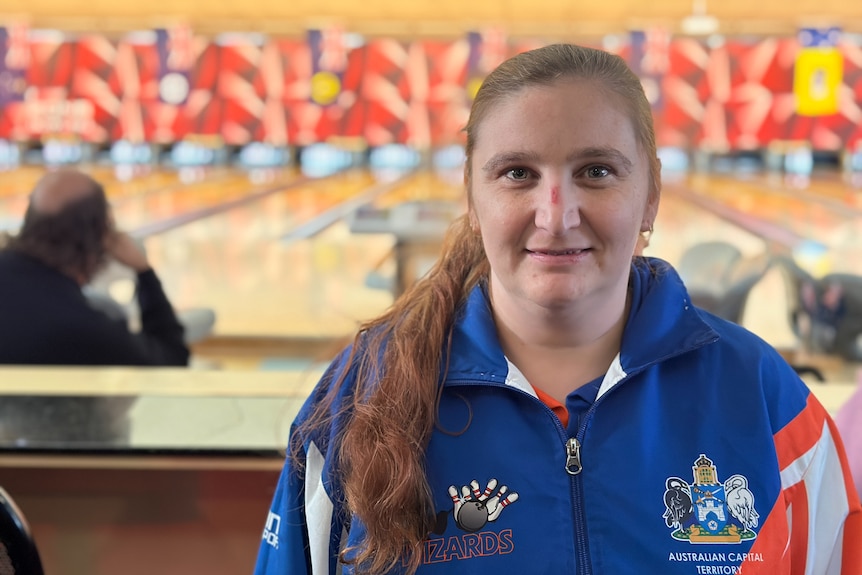 A woman smiles in a bowling alley, wearing an ACT Wizards jacket.