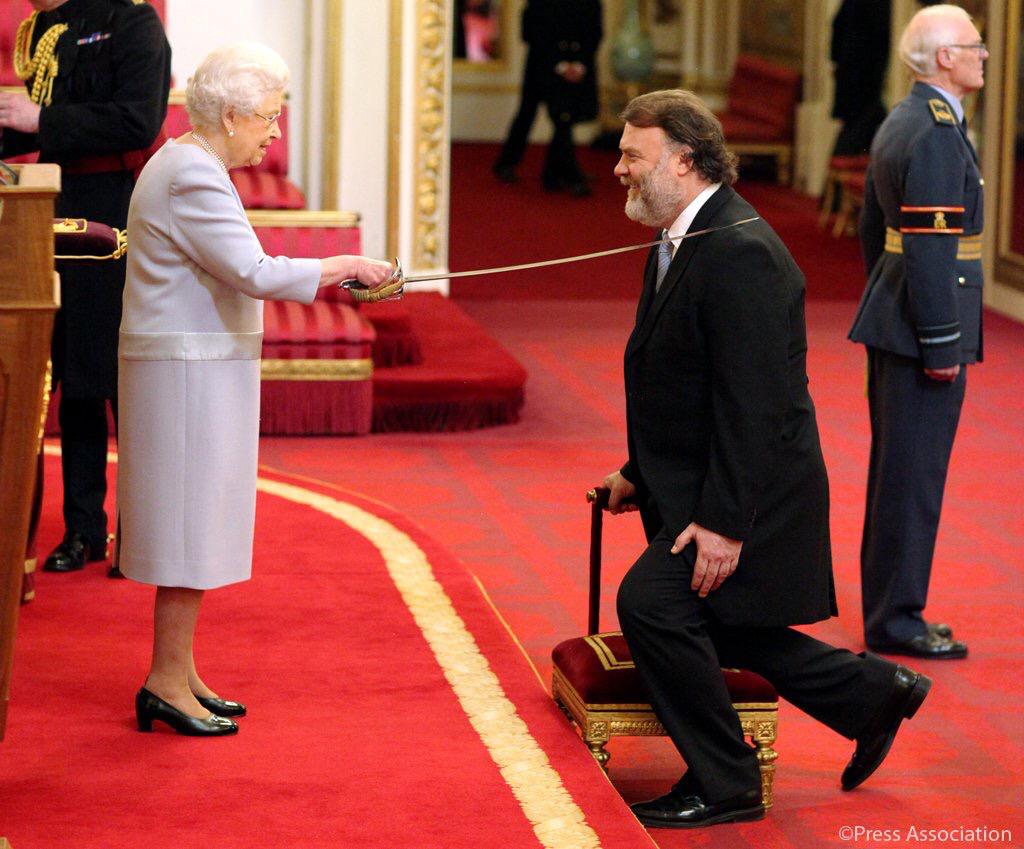Bryn Terfel kneels during an investiture ceremony. The queen touches his shoulder with a ceremonial sword.