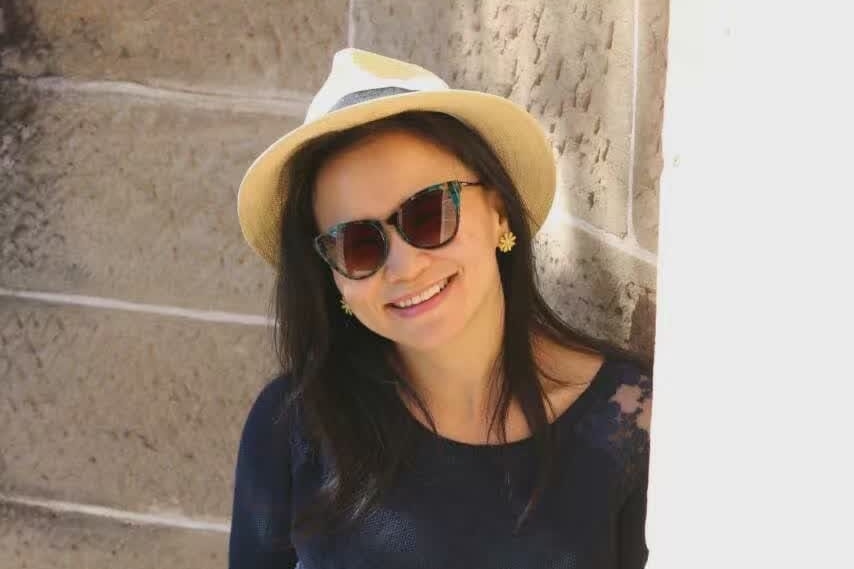 Cheng Lei leans on a wall smiling. She is wearing a hat and sunglasses.