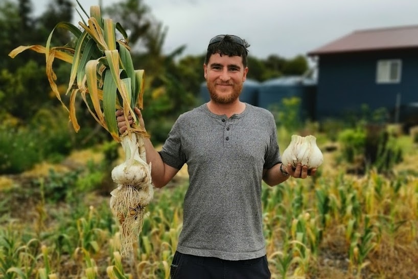 Bearded young man holding up garlic bulbs in left hand and garlic plant with right in garden, house in background.