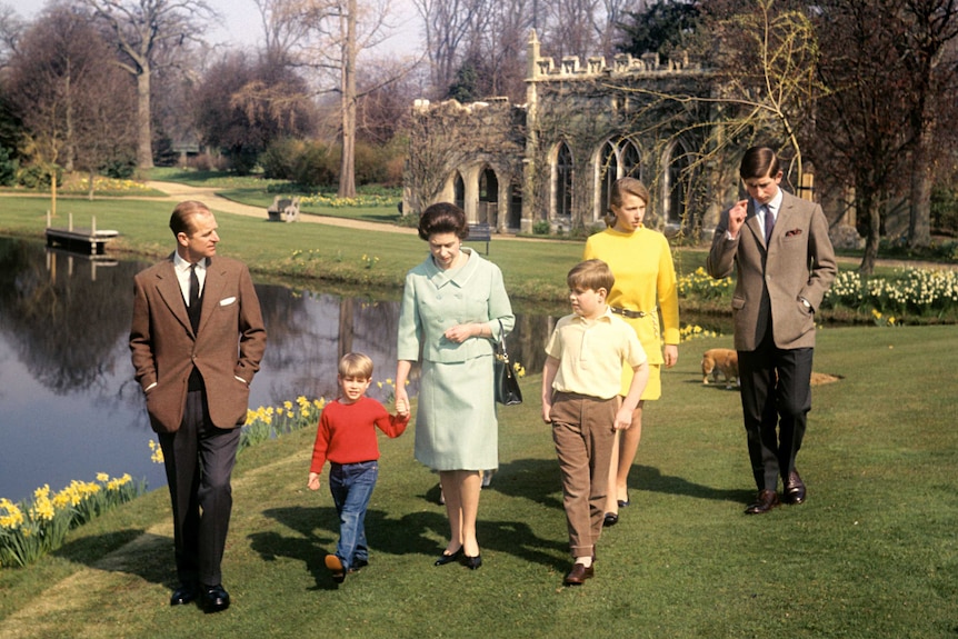 An colour film photograph shows Queen Elizabeth II with a teenage Prince Charles and family walking across verdant green lawns.