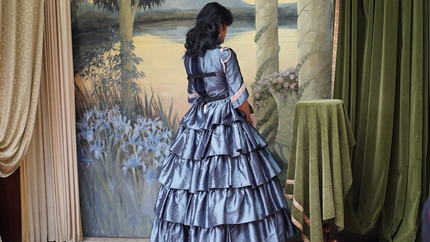 A woman stands with her back to the camera, wearing a long, full, blue dress from the 1850s