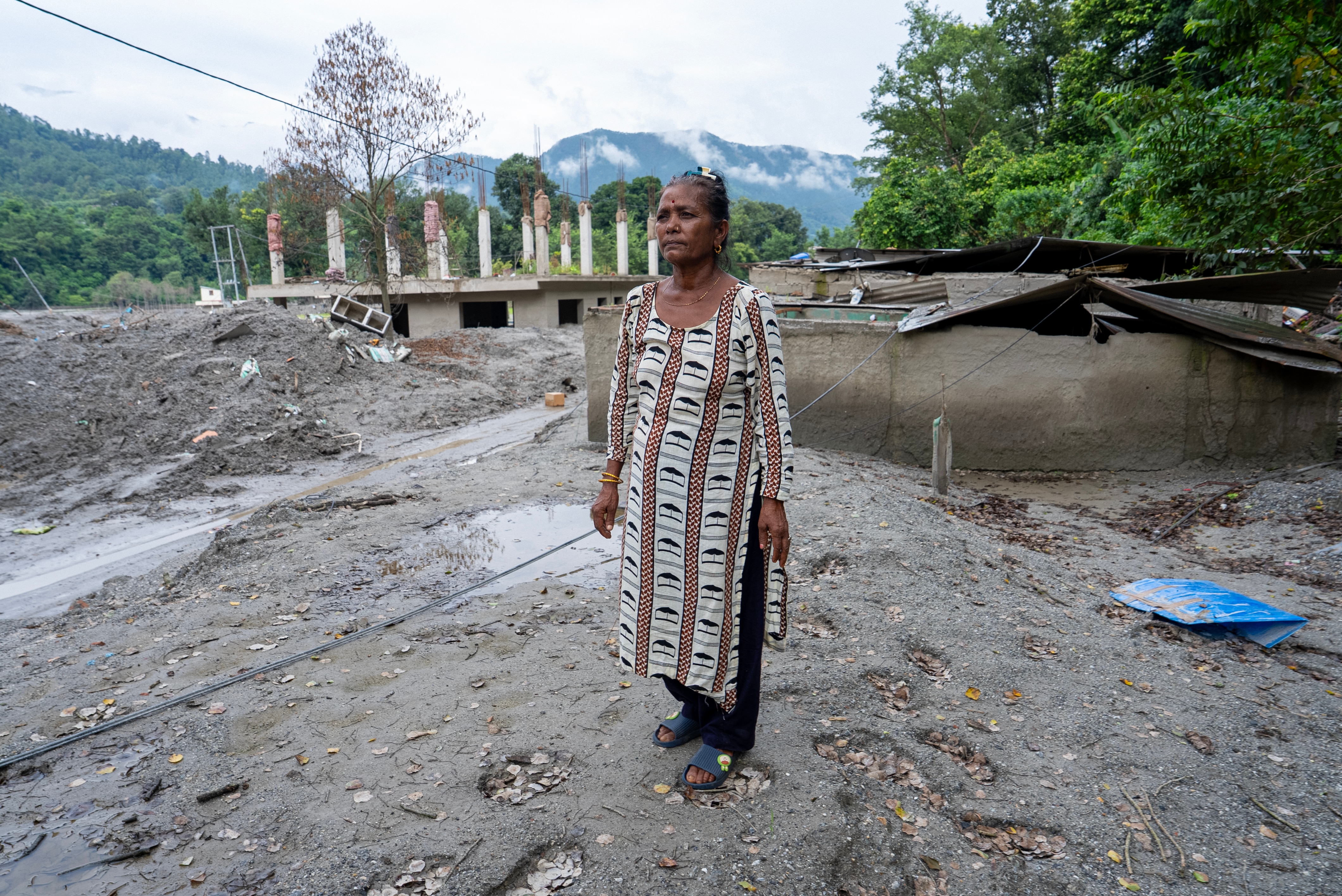 A middle aged woman dressed in a simple but elegant gown stands in a field of mud, looking longingly for the house that is no lh