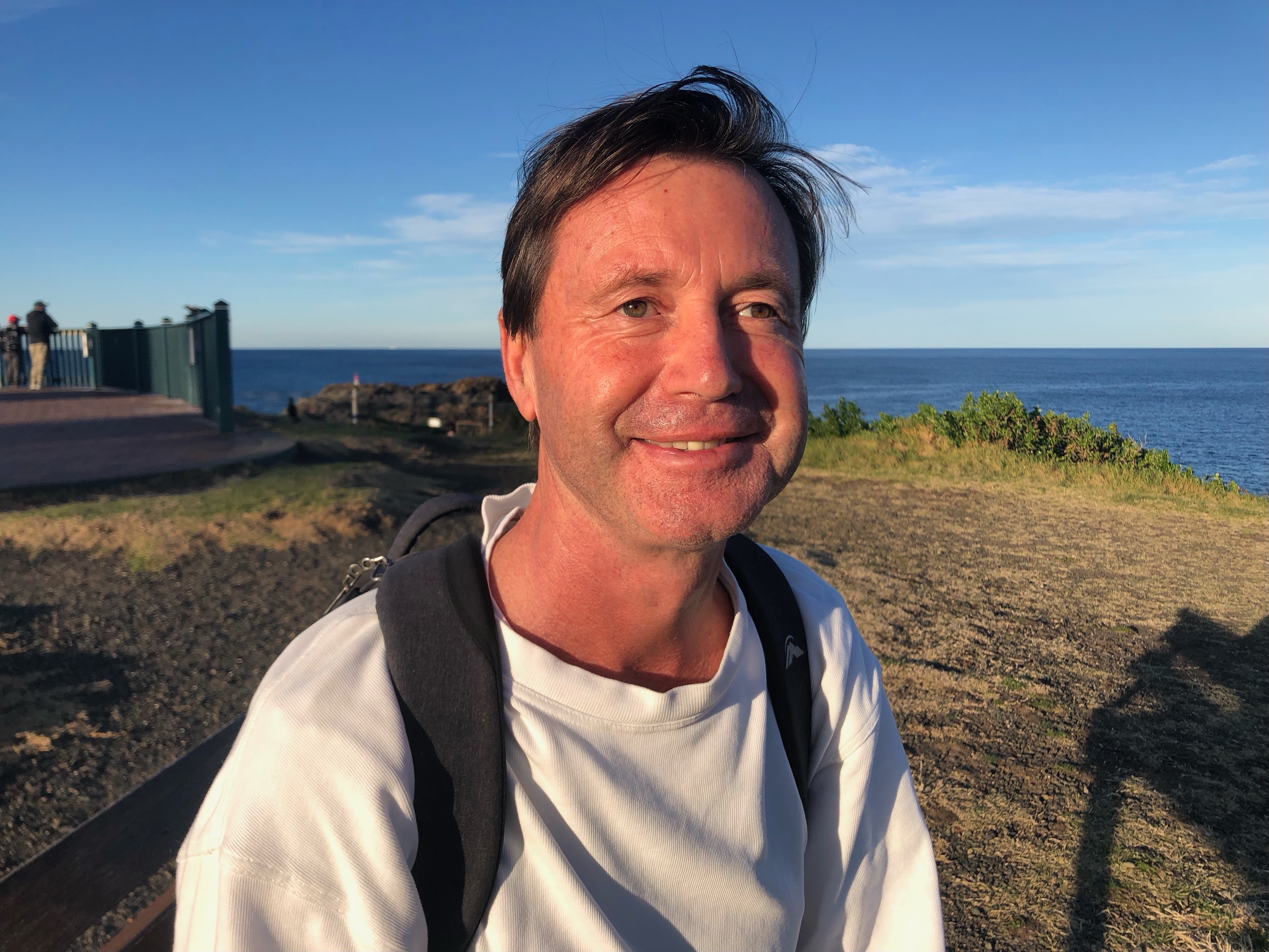 A visually impaired man stands with a backpack on, with some sand and some grass behind him.