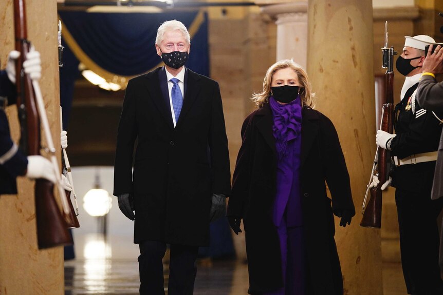 A man wearing a suit and blue tie stands next to a woman wearing a purple pantsuit, both wearing masks.
