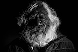 A darkly lit potrait of an Indigenous man with grey hair and beard.