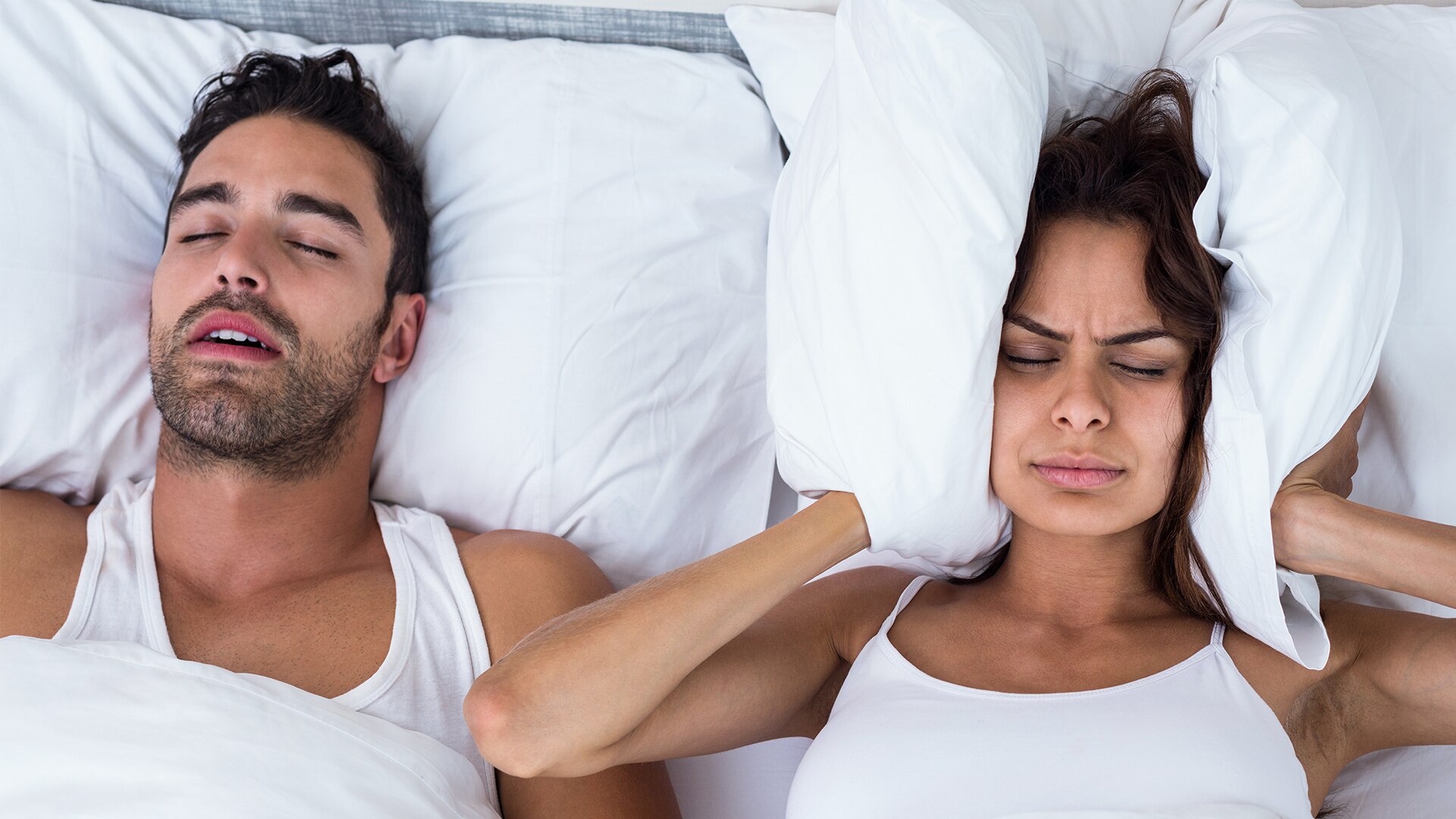 A man and woman are lying next to each other. The man is asleep while the woman has a pillow around her head covering her ears