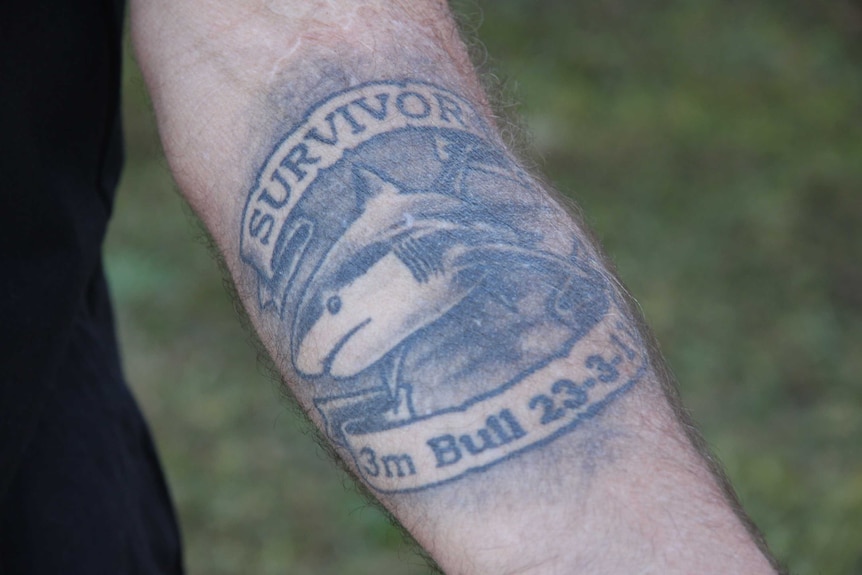 A closeup photo of a greyscale tattoo on the inside of a forearm. The tattoo depicts a shark underneath text saying "survivor".