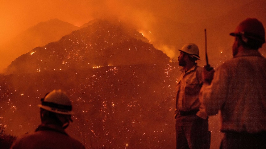 Three firefighters look out over a burning mountain. The sky and scene is completely orange from the flames and smoke.