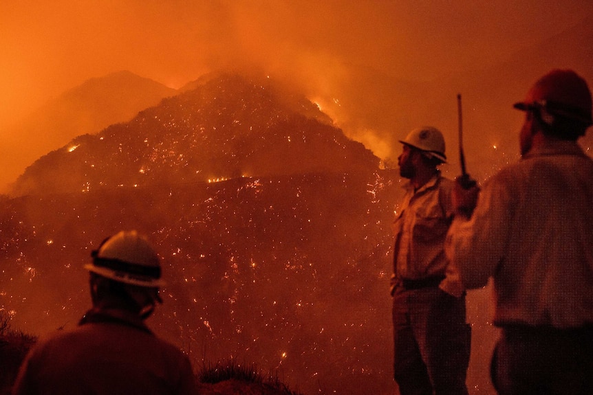 Three firefighters look out over a burning mountain. The sky and scene is completely orange from the flames and smoke.