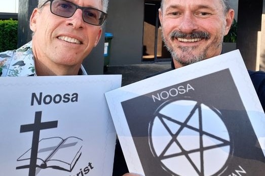 Two men with grey hair, one with glasses, holding two Noosa signs - one with a Christian Cross and the other with pentagram