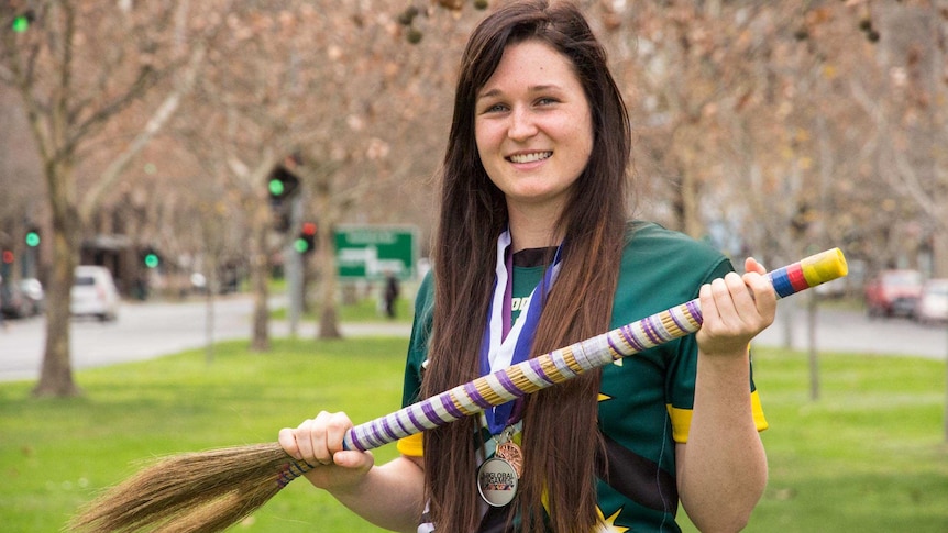 A woman dressed in Australian sporting colours and wearing medals holds a broomstick