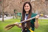A woman dressed in Australian sporting colours and wearing medals holds a broomstick