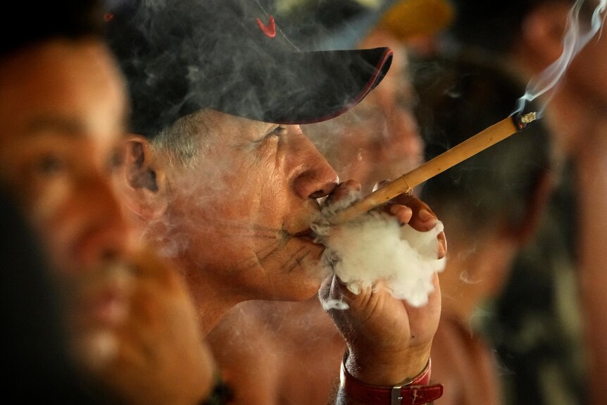 A man in a black cap smokes a cigar, exhaling a cloud of smoke around it 