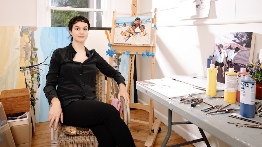 Portrait of Thea Anamara Perkins, wearing black and sitting in a chair amongst her work and an artists table
