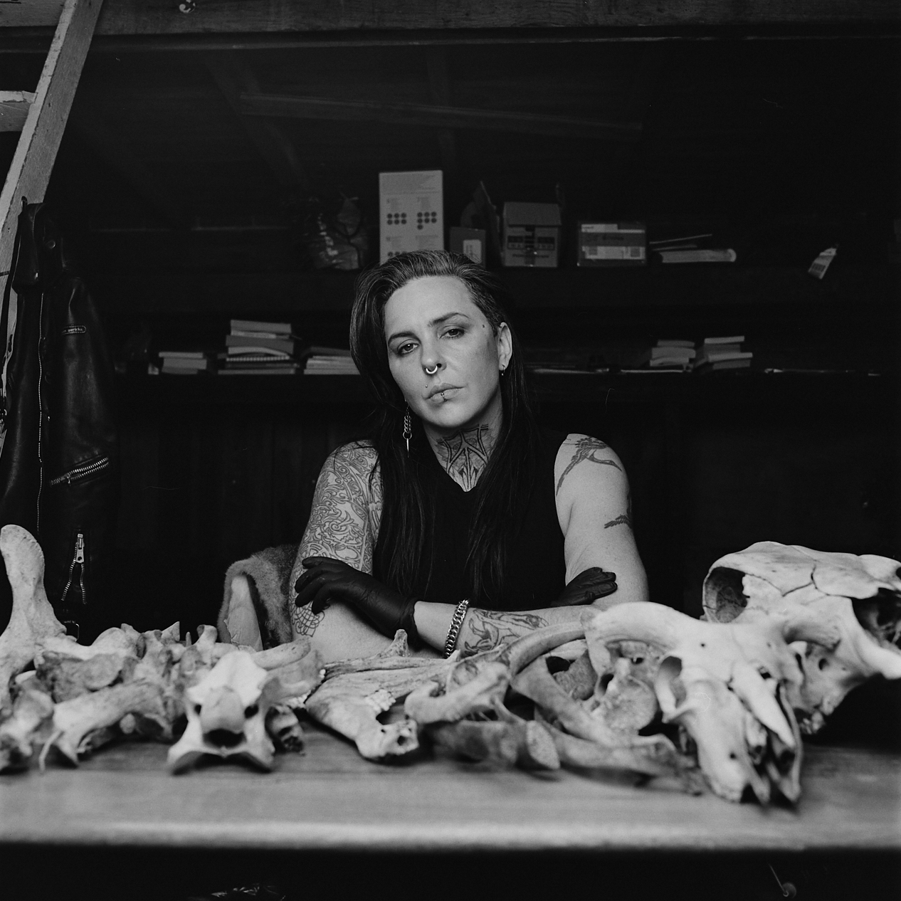 A B&W image of a young person with long hair sitting behind a collection of animal bones arranged in a row on a wooden bench 